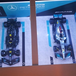 K&ouml;nnte beinhalten: Zwei Formel-1-Rennwagen, einer schwarz und einer silber, werden auf einem blauen und wei&szlig;en Hintergrund ausgestellt. Die Autos geh&ouml;ren beide zum Petronas Formel-1-Team. Das schwarze Auto tr&auml;gt die Nummer 44. Das silberne Auto tr&auml;gt das AMG Petronas F1 Team Logo.