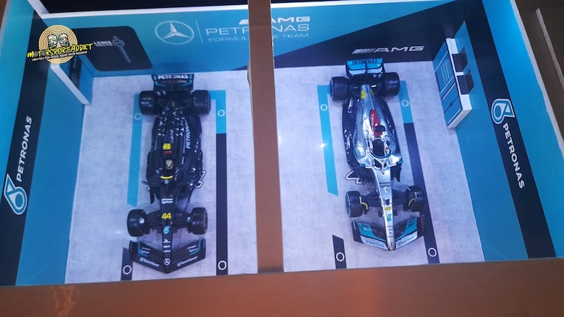 K&ouml;nnte beinhalten: Zwei Formel-1-Rennwagen, einer schwarz und einer silber, werden auf einem blauen und wei&szlig;en Hintergrund ausgestellt. Die Autos geh&ouml;ren beide zum Petronas Formel-1-Team. Das schwarze Auto tr&auml;gt die Nummer 44. Das silberne Auto tr&auml;gt das AMG Petronas F1 Team Logo.