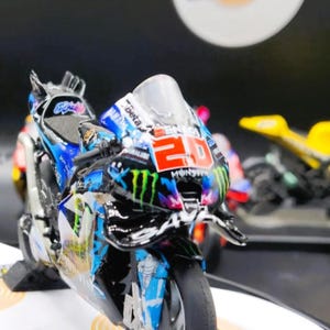 Peut inclure: Modèle réduit de moto détaillé, principalement noir et bleu, avec le numéro 20 bien en évidence. Le modèle présente des logos de sponsors et un pare-brise transparent. La moto est posée sur une surface blanche avec des motifs circulaires.