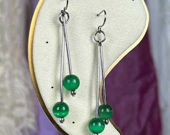 Puede incluir: Un par de pendientes colgantes con hilos de metal plateado y cuentas redondas de color verde vibrante. Cada pendiente consta de dos varillas de metal delgadas suspendidas de un gancho, con cuentas verdes unidas a diferentes longitudes para crear un efecto.