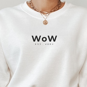 May include: White crewneck sweatshirt with the text "WOW EST. 2001" printed on the front. The sweatshirt is being worn by a person wearing blue jeans.