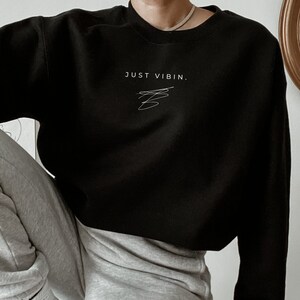 May include: A black cropped sweatshirt with the text "JUST VIBIN." in white. The model is wearing the sweatshirt with grey sweatpants and white socks.