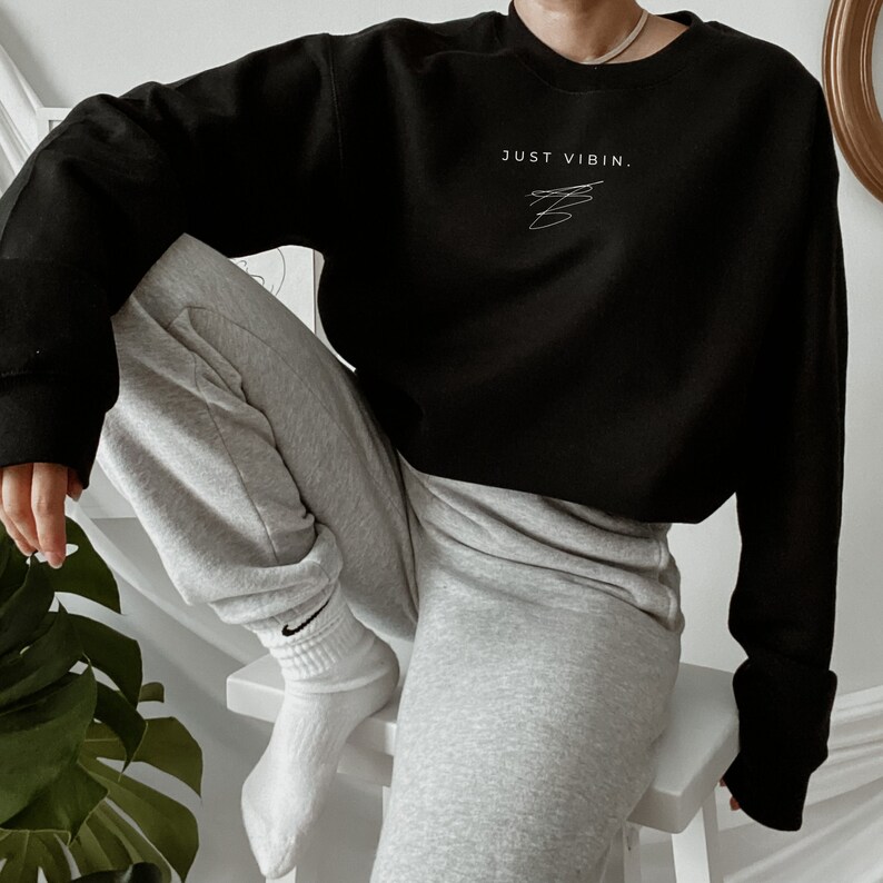 May include: A black cropped sweatshirt with the text "JUST VIBIN." in white. The model is wearing the sweatshirt with grey sweatpants and white socks.