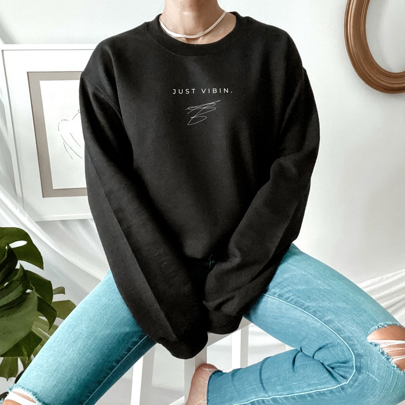 May include: Black sweatshirt with the text "JUST VIBIN." in white lettering. The sweatshirt is worn by a person wearing blue jeans.