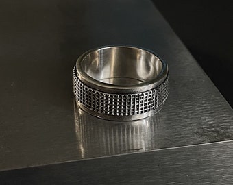 Op de afbeelding: Een zilveren ring met een getextureerd, rasterachtig patroon rond de band. De ring is gemaakt van glanzend metaal en ligt op een grijs oppervlak. De ring is een sieraad.