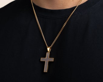 Op de afbeelding: Goudkleurige ketting met een kruisvormige hanger. Het kruis heeft een grijze kern en een gouden rand. De ketting heeft een delicate curb-link stijl. De ketting wordt gedragen over een zwart T-shirt.