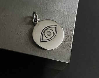 Op de afbeelding: Een kleine, ronde zilveren hanger met een gegraveerd oogontwerp. De hanger is bevestigd aan een kleine lus voor een ketting. Het oogontwerp is een eenvoudige, geometrische weergave.