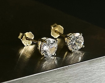 Op de afbeelding: Een paar gouden oorstekers met grote, ronde, heldere edelstenen. De oorbellen zijn gezet in goudmetaal en worden gepresenteerd op een donker, getextureerd oppervlak. De studs zijn eenvoudig en elegant.