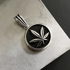 May include: A silver pendant with a cannabis leaf design set in a black circular frame. The pendant has a detailed, textured finish and hangs from a silver bail. The background is a textured, metallic surface.