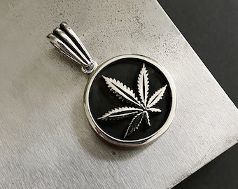 Op de afbeelding: Zilveren hanger met een cannabisbladontwerp in een zwart cirkelvormig frame. De hanger heeft een gedetailleerde, getextureerde afwerking en hangt aan een zilveren balein. De achtergrond is een getextureerd, metalen oppervlak.