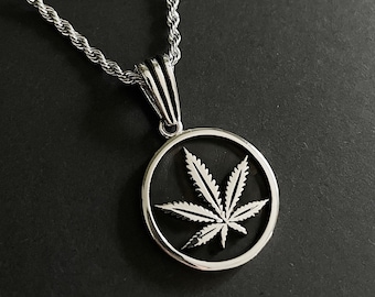 Op de afbeelding: Zilverkleurige ketting met een ronde hanger met een cannabisblad. De hanger hangt aan een touwketting. De ketting wordt getoond tegen een donkere achtergrond.