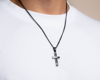 Op de afbeelding: Een zwarte kruis hanger ketting. Het kruis is zilverkleurig met een zwarte ketting. De ketting wordt gedragen over een wit t-shirt. De achtergrond is neutraal.