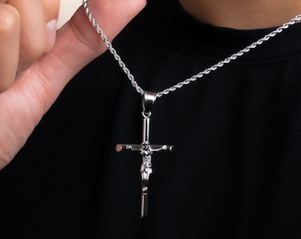 Op de afbeelding: Zilverkleurige kruis-hangerketting. De hanger toont een gedetailleerde afbeelding van Jezus aan het kruis. De ketting heeft een fijne ketting. De achtergrond is effen zwart.