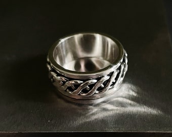 Op de afbeelding: Een zilverkleurige ring met een kettinglink-ontwerp rond de band. De ring staat tegen een donkere achtergrond, waardoor de metalen glans wordt benadrukt. Het ontwerp van de ring heeft een reeks onderling verbonden schakels.