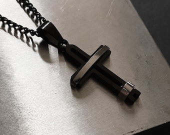 Op de afbeelding: Een zwarte kruis-hangerketting met een bijpassende ketting. Het kruis heeft een gelaagd ontwerp met een zilverkleurig accent. De ketting wordt gepresenteerd op een getextureerd, metalen oppervlak.