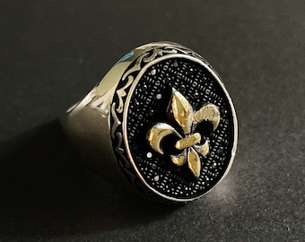 Op de afbeelding: Zilveren ring met een zwarte voorkant met een gouden fleur-de-lis embleem. De ring heeft een sierlijke, patroonrand en een gepolijste afwerking. De fleur-de-lis is een symbool van koninklijke macht.