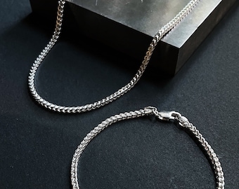 May include: Two silver chain necklaces on a black background. The larger chain is laying on a black rectangular surface. The smaller chain is laying on the black background.