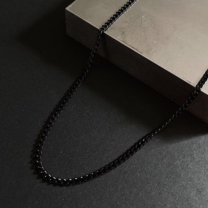 Peut inclure: Un collier chaîne noir sur une surface grise. La chaîne est en métal et a un design à maillons plats.