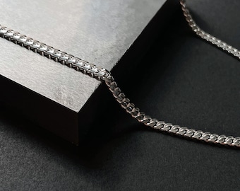 Op de afbeelding: Een zilveren gourmetketting die wordt getoond op een grijze, schuine ondergrond tegen een zwarte achtergrond. De ketting bestaat uit in elkaar grijpende, afgeplatte schakels die licht reflecteren. De sieraden zijn een eenvoudig, maar elegant accessoire.