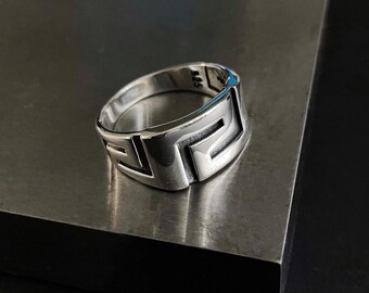 Op de afbeelding: Een zilveren ring met een geometrisch ontwerp. De ring heeft een reeks rechthoekige vormen en lijnen, waardoor een moderne en minimalistische esthetiek ontstaat. De ring wordt gepresenteerd op een metalen oppervlak, wat de gepolijste afwerking benadrukt.
