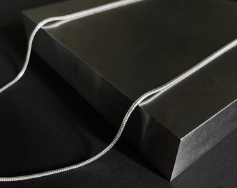 Op de afbeelding: Een zilveren slangencolier rust op een donkergrijze, rechthoekige blok. De ketting heeft een glad, reflecterend oppervlak en het blok heeft een geborsteld metalen uiterlijk. De achtergrond is effen zwart, wat een contrastrijk beeld creëert.