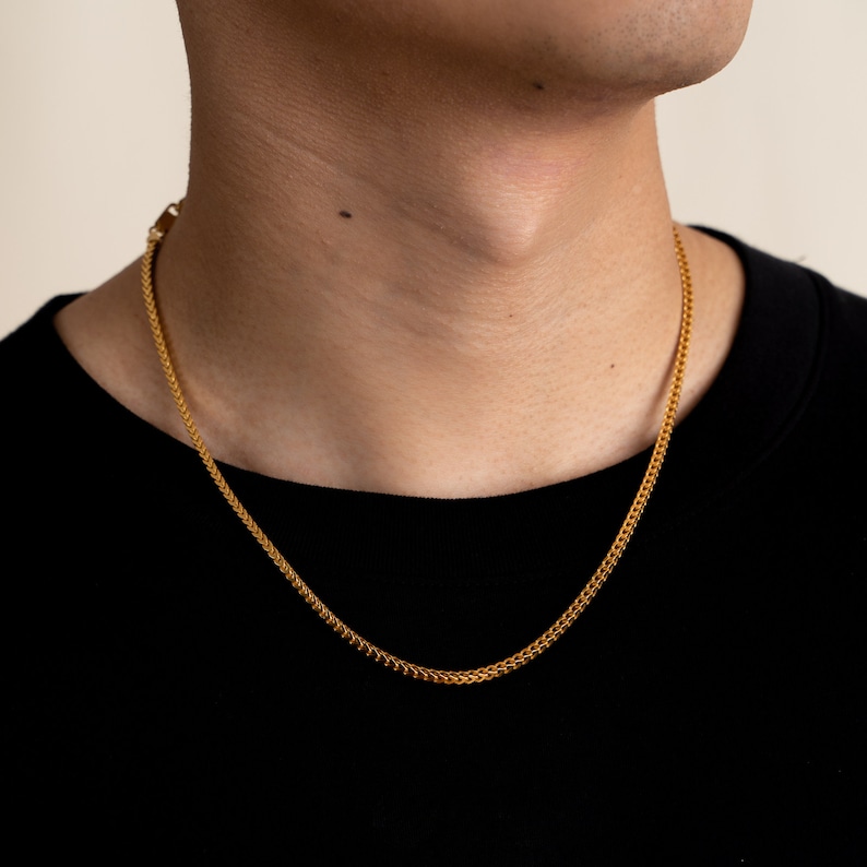 K&ouml;nnte beinhalten: Eine goldene Kettenkette mit einem zarten, filigranen Design. Die Kette wird von einer Person mit einem dunklen Hemd getragen.