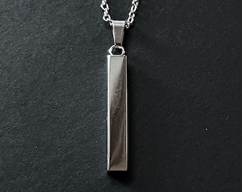Op de afbeelding: Een zilverkleurige hangerketting met een rechthoekige hanger. De hanger hangt aan een fijne ketting tegen een donkere achtergrond. De ketting is gemaakt van glanzend, reflecterend metaal.