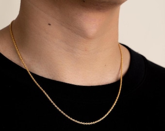 Op de afbeelding: Een goudkleurige touwketting. De ketting is om de nek van een persoon gedrapeerd, tegen een zwart shirt. De ketting heeft een gedraaid touwontwerp en is helder goudkleurig.