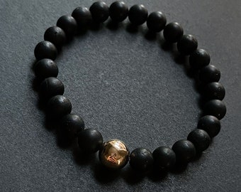 Op de afbeelding: Zwart kralenarmband met één goudkleurige kraal. De armband bestaat uit matte zwarte, ronde kralen. De gouden kraal is glad en reflecterend, wat een contrast creëert.