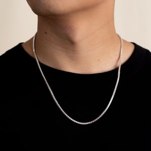 Op de afbeelding: Een zilveren kettingketting die om de nek van een persoon wordt gedragen. De ketting is in box-kettingstijl.