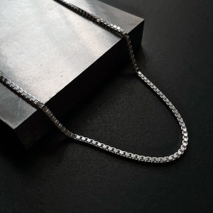 Op de afbeelding: Een zilveren ketting met een box chain-ontwerp. De ketting ligt op een grijs oppervlak.