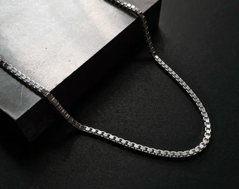 May include: A silver chain necklace with a box chain design. The chain is laying on a gray surface.
