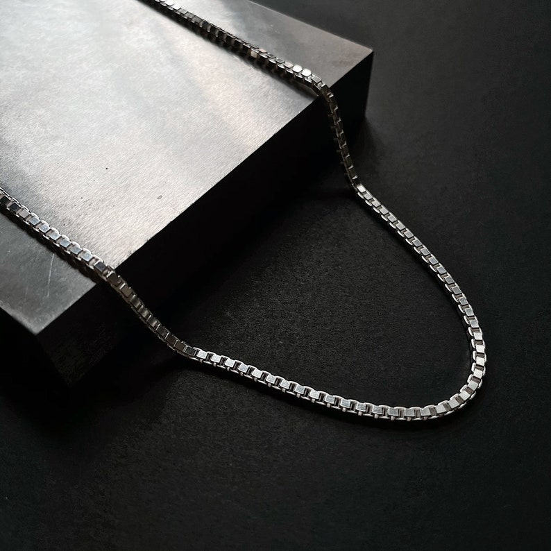 Op de afbeelding: Een zilveren ketting met een box chain-ontwerp. De ketting ligt op een grijs oppervlak.