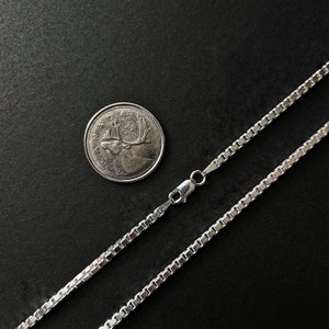 Op de afbeelding: Een zilveren ketting met een karabijn sluiting. De ketting is van het box-type en ligt op een zwarte ondergrond naast een Canadese munt van 25 cent.