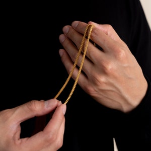 K&ouml;nnte beinhalten: Eine goldene Kettenkette mit einem zarten, filigranen Design. Die Kette wird in zwei H&auml;nden gehalten, wodurch ihre L&auml;nge und Details zur Geltung kommen.