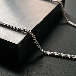 Op de afbeelding: Een zilveren ketting met een box chain-ontwerp. De ketting ligt op een grijs oppervlak.