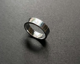Op de afbeelding: Een glanzende, zilveren ring met een platte band. De ring is gemaakt van een reflecterend metaal en het oppervlak is glad. De ring wordt tegen een donkere achtergrond getoond. De ring is een sieraad.