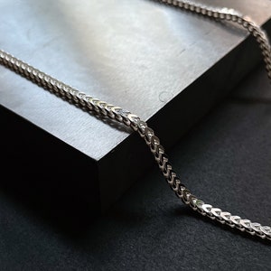 May include: A silver chain necklace with a curb link pattern. The chain is lying on a gray surface.