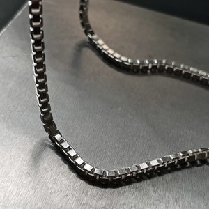 Op de afbeelding: Een zilveren ketting met een box chain ontwerp. De ketting ligt op een grijs oppervlak.