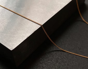 Op de afbeelding: Een fijne gouden ketting ligt over een blok met textuur in zilverkleur. De fijne schakels van de ketting vangen het licht op en creëren een subtiele glinstering tegen de donkere achtergrond. De ketting lijkt een minimalistisch sieraad te zijn.