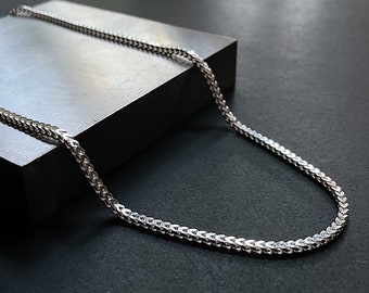 May include: A silver chain necklace with a curb link style. The chain is laying on a gray surface.