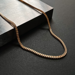 K&ouml;nnte beinhalten: Eine goldene Kettenkette im Curb-Link-Stil. Die Kette liegt auf einer grauen Oberfl&auml;che.