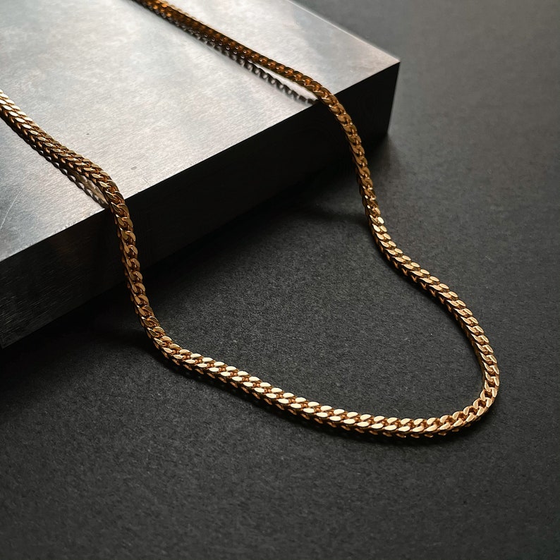 K&ouml;nnte beinhalten: Eine goldene Kettenkette im Curb-Link-Stil. Die Kette liegt auf einer grauen Oberfl&auml;che.