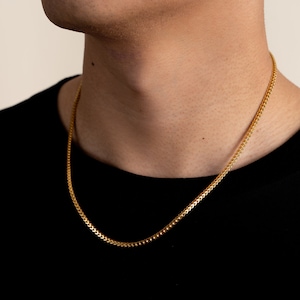 K&ouml;nnte beinhalten: Eine goldene Kettenkette mit einem filigranen, komplizierten Design. Die Kette wird von einer Person mit einem schwarzen Hemd getragen.