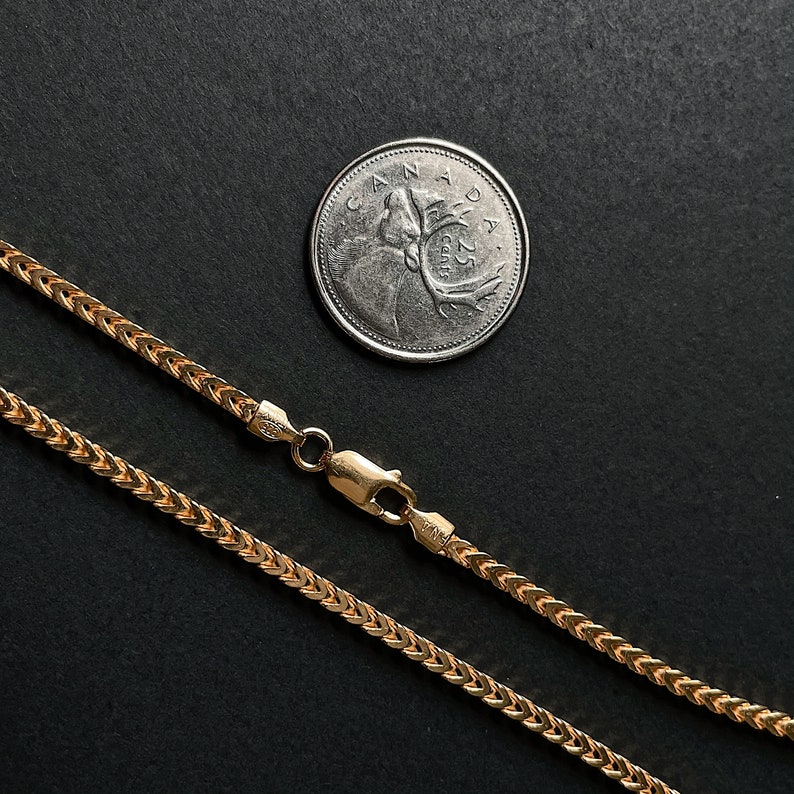 K&ouml;nnte beinhalten: Eine goldene Kettenkette mit einem Weizenkettenmuster. Die Kette liegt auf einer schwarzen Oberfl&auml;che neben einer kanadischen 25-Cent-M&uuml;nze.