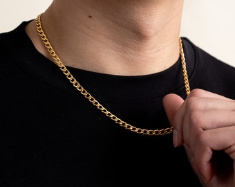 Op de afbeelding: Een goudkleurige gourmette ketting. De ketting is gemaakt van kleine, in elkaar grijpende schakels en wordt om de nek gedragen. De persoon draagt een zwart t-shirt.