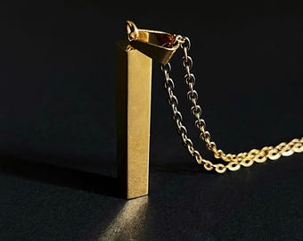 Op de afbeelding: Een goudkleurige ketting met een geometrische, rechthoekige hanger. De hanger hangt aan een fijne ketting. De ketting is geplaatst tegen een donkere achtergrond. De hanger heeft een gepolijste afwerking.