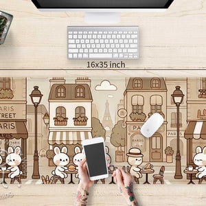 Op de afbeelding: Een bruine en witte muismat met een cartoonillustratie van een Parijse straat. De scène bevat gebouwen, lantaarns en cartoondieren. De muismat is 16 inch bij 35 inch.