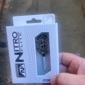 Op de afbeelding: Een witte kartonnen doos met een set dartflights en shafts. De verpakking bevat de tekst Nitro Flite en Intermediate No. 2 28mm. De dartflight is zwart met een rond Stone Island-logo met een kompasontwerp in het midden.