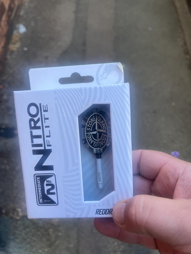 Może przedstawiać: Białe kartonowe pudełko zawierające zestaw pi&oacute;rek i shaft&oacute;w do rzutek. Opakowanie zawiera napis Nitro Flite oraz Intermediate No. 2 28mm. Pi&oacute;rko jest czarne z okrągłym logo Stone Island z motywem kompasu umieszczonym w centrum.